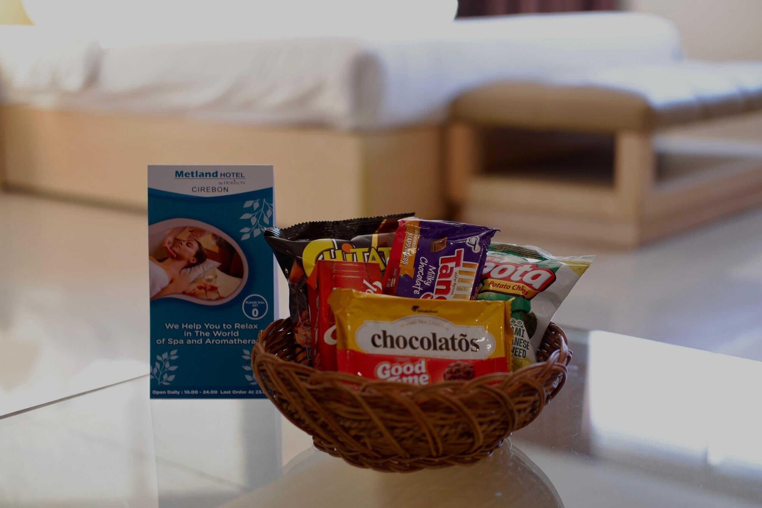 Habitación de hotel con una cesta de snacks que incluye chocolate y papas fritas, un letrero del Hotel Metland Cirebon y una cama con ropa blanca.