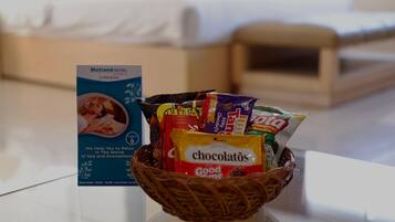 Habitación de hotel con una cesta de snacks que incluye chocolate y papas fritas, un letrero del Hotel Metland Cirebon y una cama con ropa blanca.