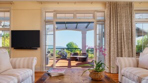 Uma sala de estar com uma grande mesa de centro de madeira, dois sofás listrados, uma TV fixada na parede e vista para um pátio externo com pérgola, cadeiras e um vaso de planta.