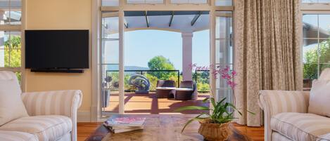 A living room with a large wooden coffee table, two striped sofas, a TV mounted on the wall, and a view of an outdoor patio with a pergola, chairs, and a potted plant.