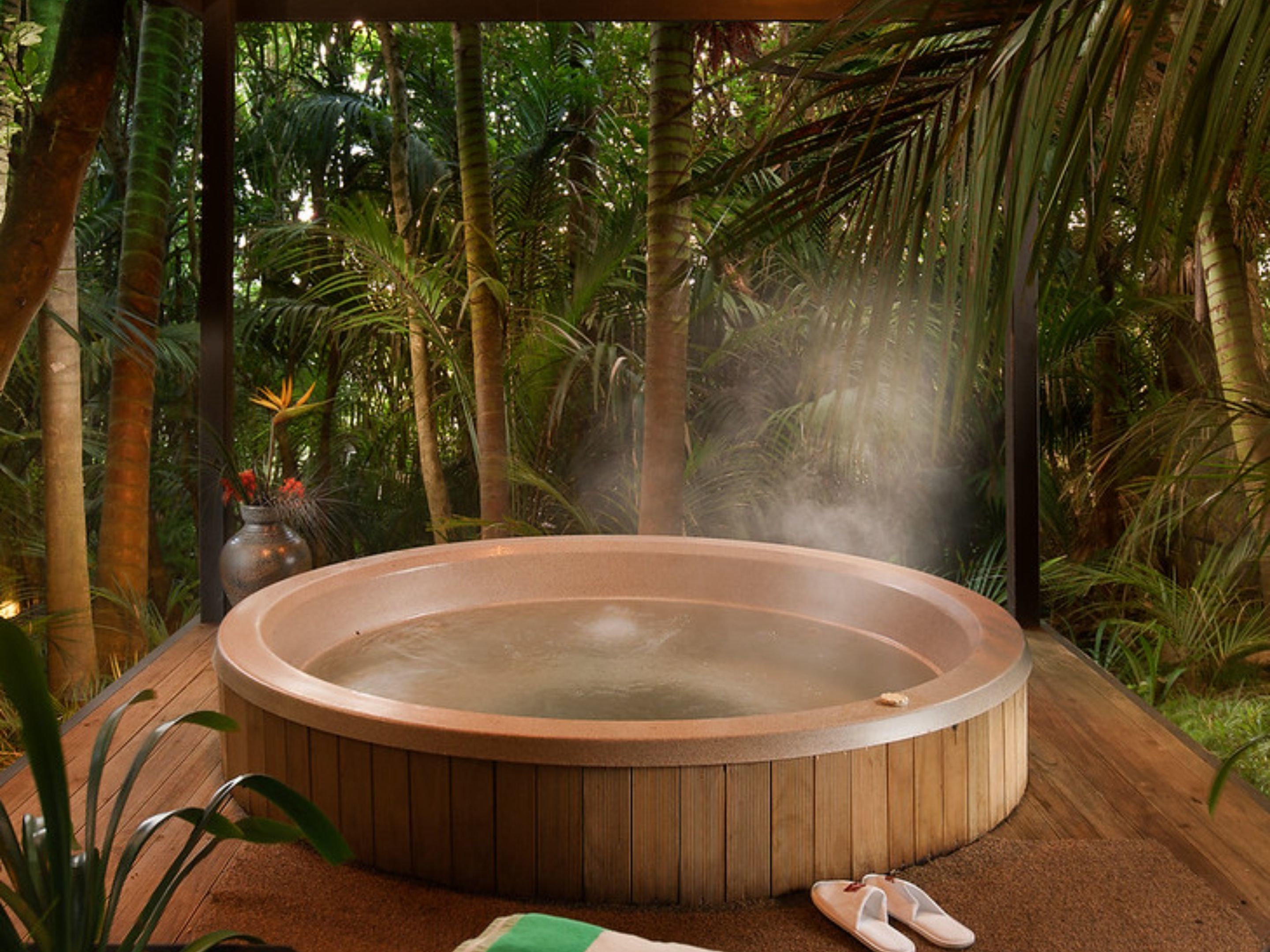 A hot tub surrounded by lush greenery, with a wooden deck and a small table with a vase and flowers.