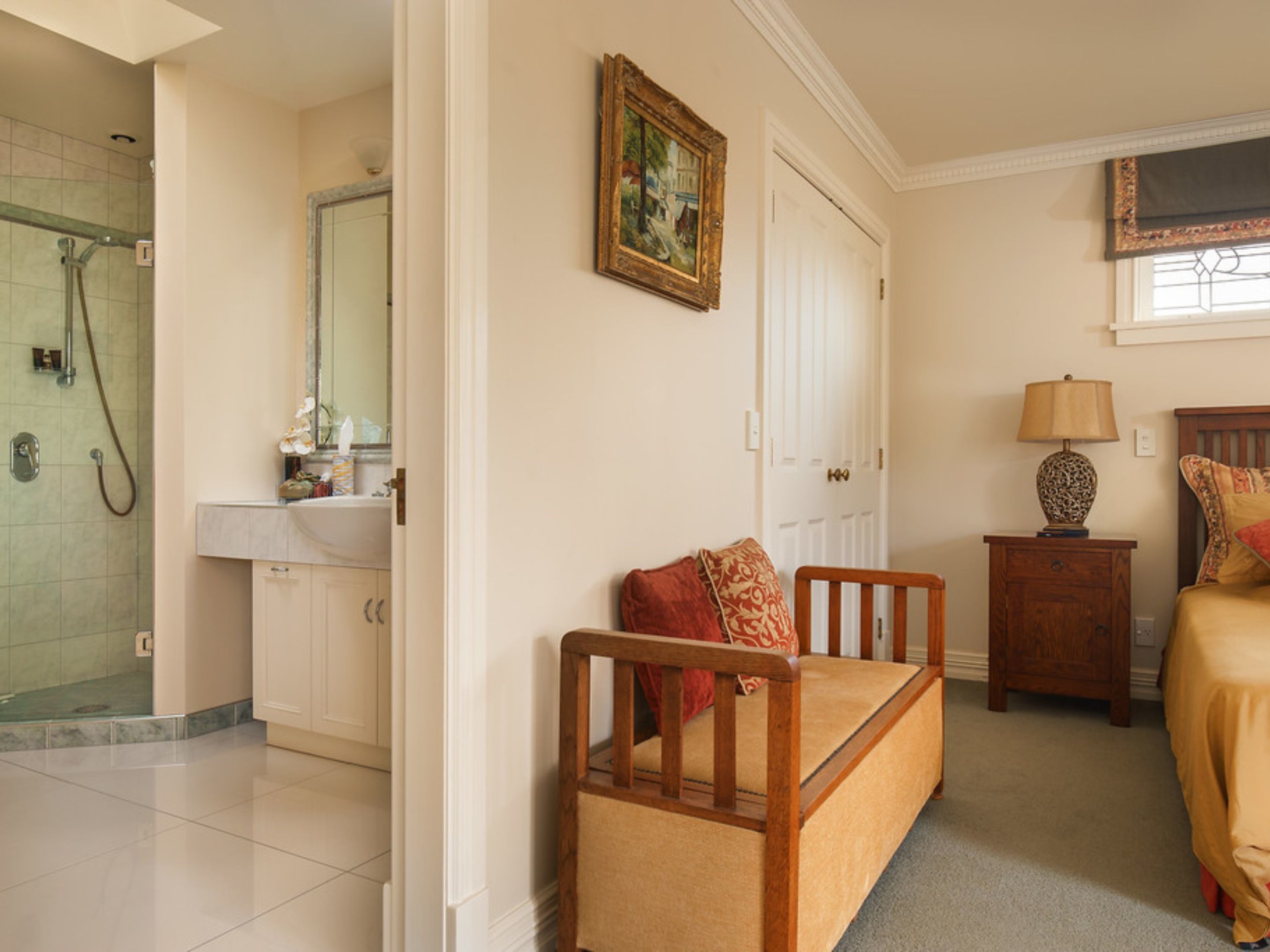 A bedroom with a bed, a nightstand, a lamp, a wooden chair, a door, a bathroom with a shower, and a mirror.