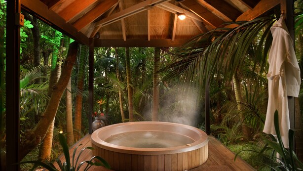 Uma banheira de hidromassagem em cenário tropical, com vigas de madeira, rodeada por vegetação exuberante.