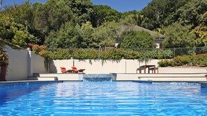 Piscina com água azul cristalina, cercada por um muro branco e um jardim com plantas e um banco.