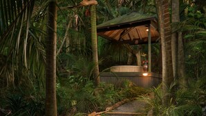 Uma área externa coberta com um caminho de pedrinhas que leva a uma estrutura de madeira, cercada por vegetação tropical densa.
