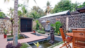 A patio with a stone wall, a statue, a small pond, and wooden furniture.