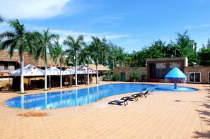 A swimming pool with lounge chairs and umbrellas, surrounded by palm trees and a building with a tiled roof. - New Wave Vung Tau (Ho Chi Minh City)