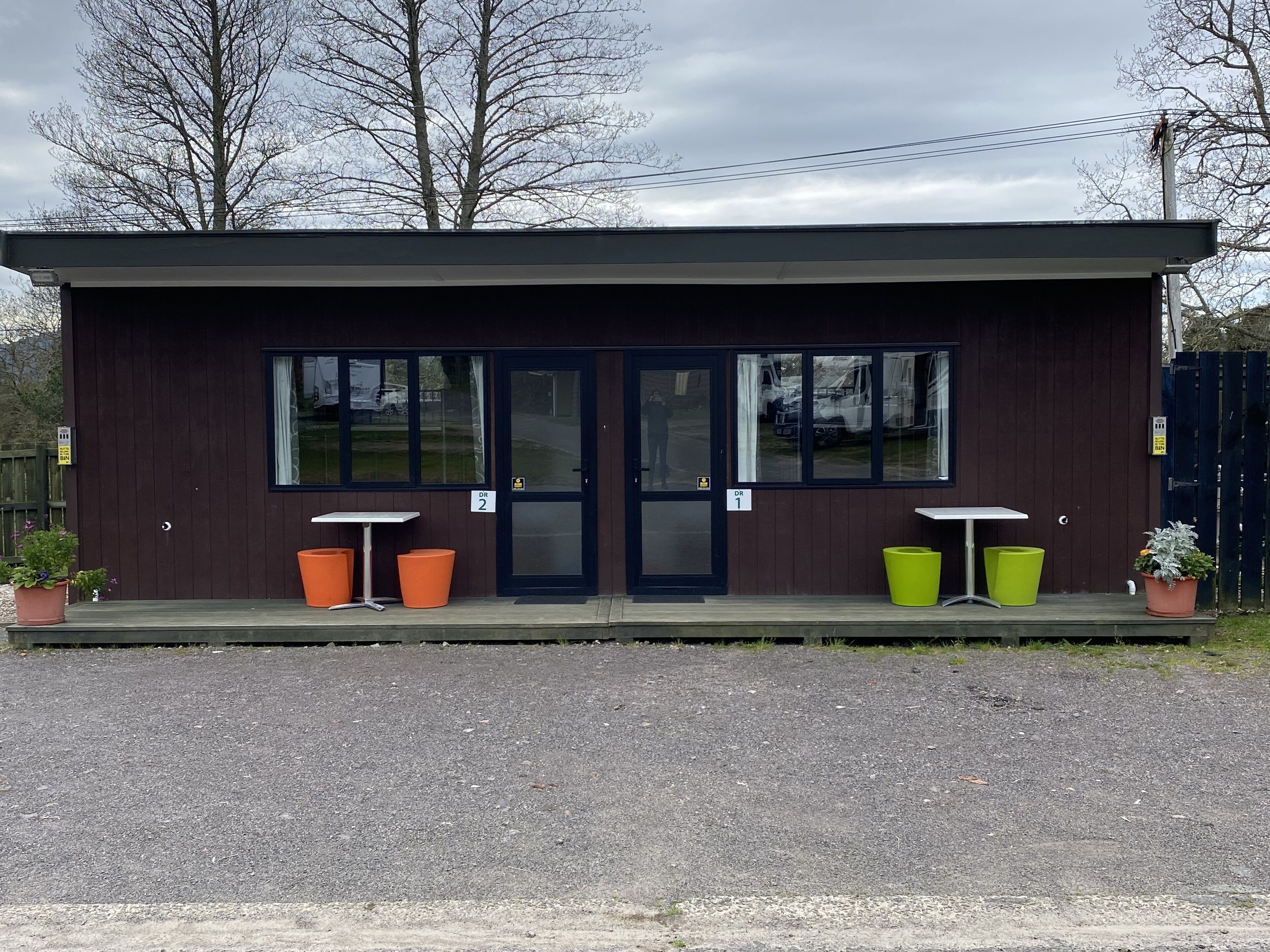 A small, two-room building with a dark brown exterior, numbered doors, and potted plants.