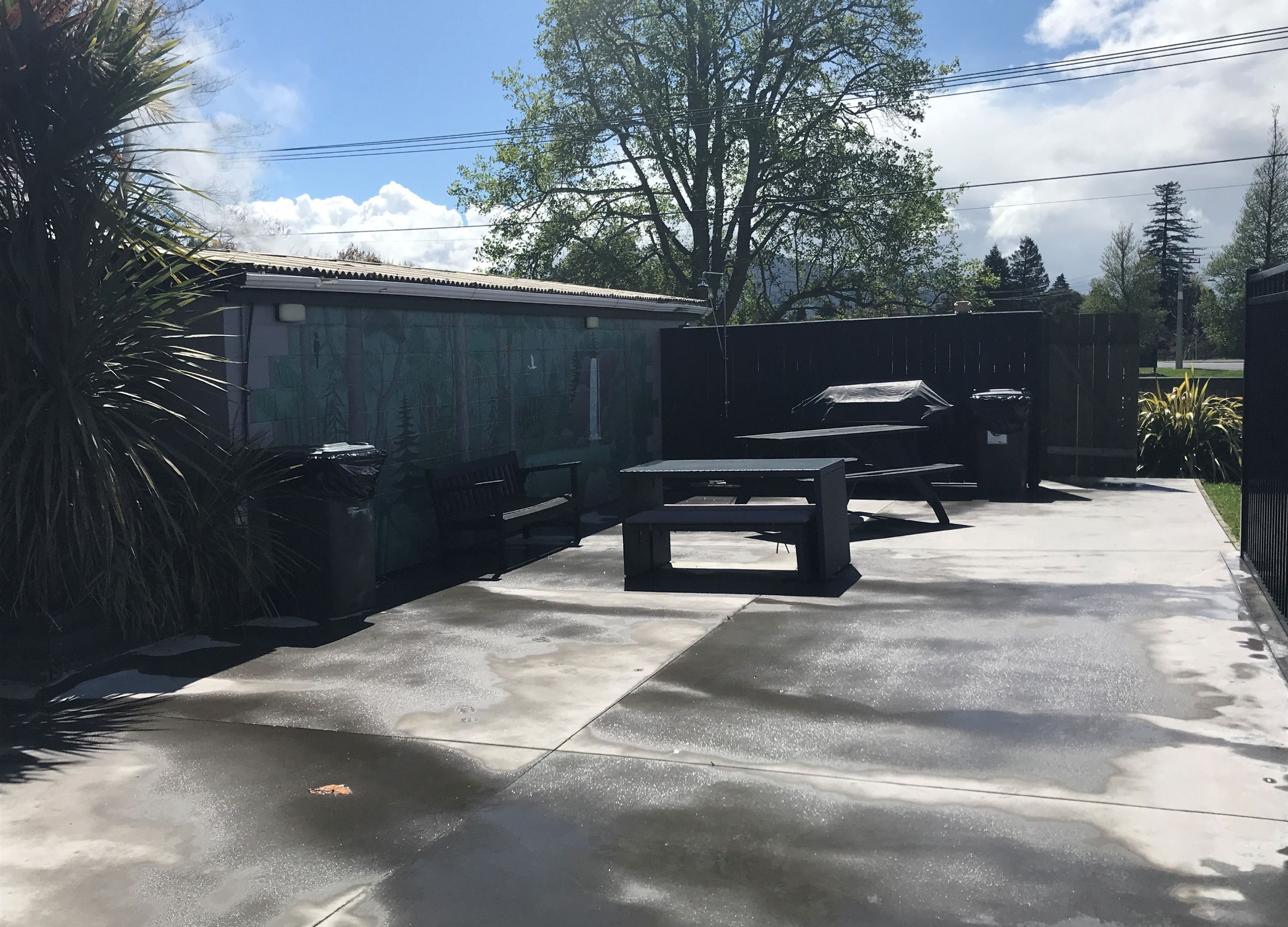 A concrete patio with picnic tables and benches, surrounded by a fence and trees.