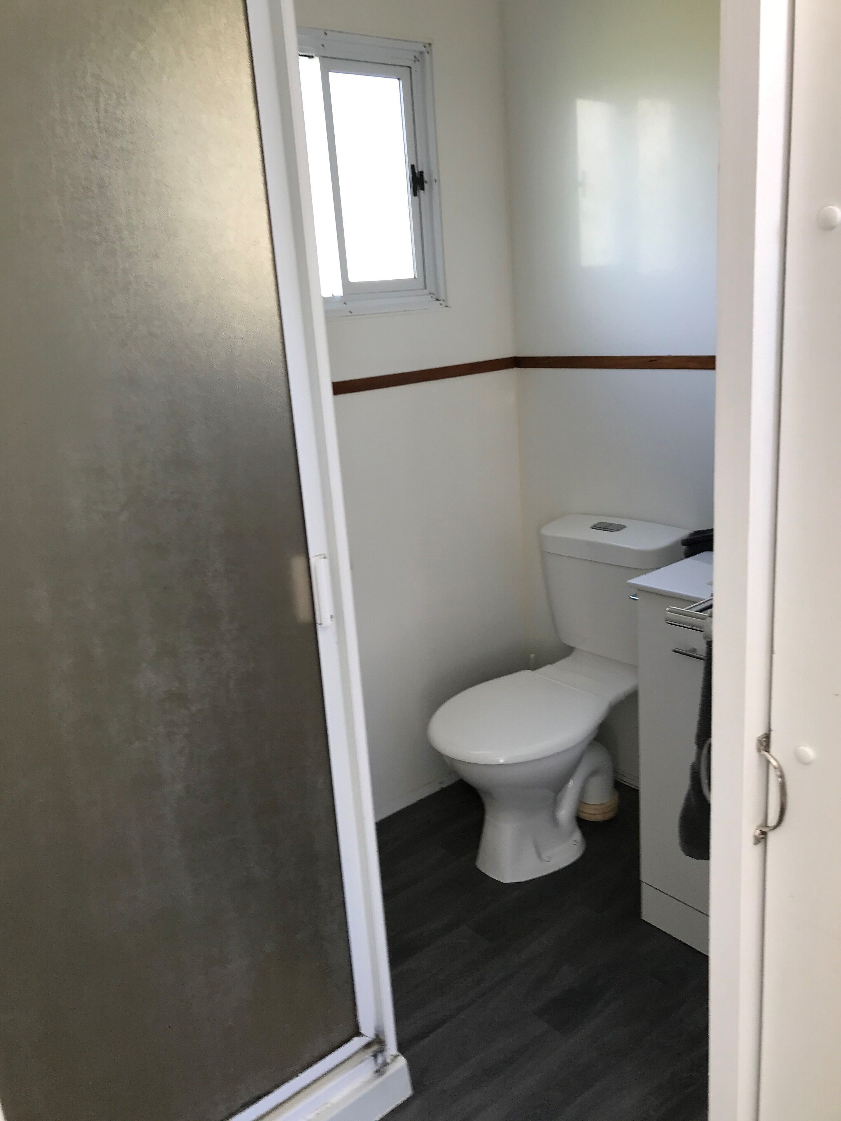 A small, compact bathroom with a white toilet, a small window above it, and a wooden trim along the lower wall.