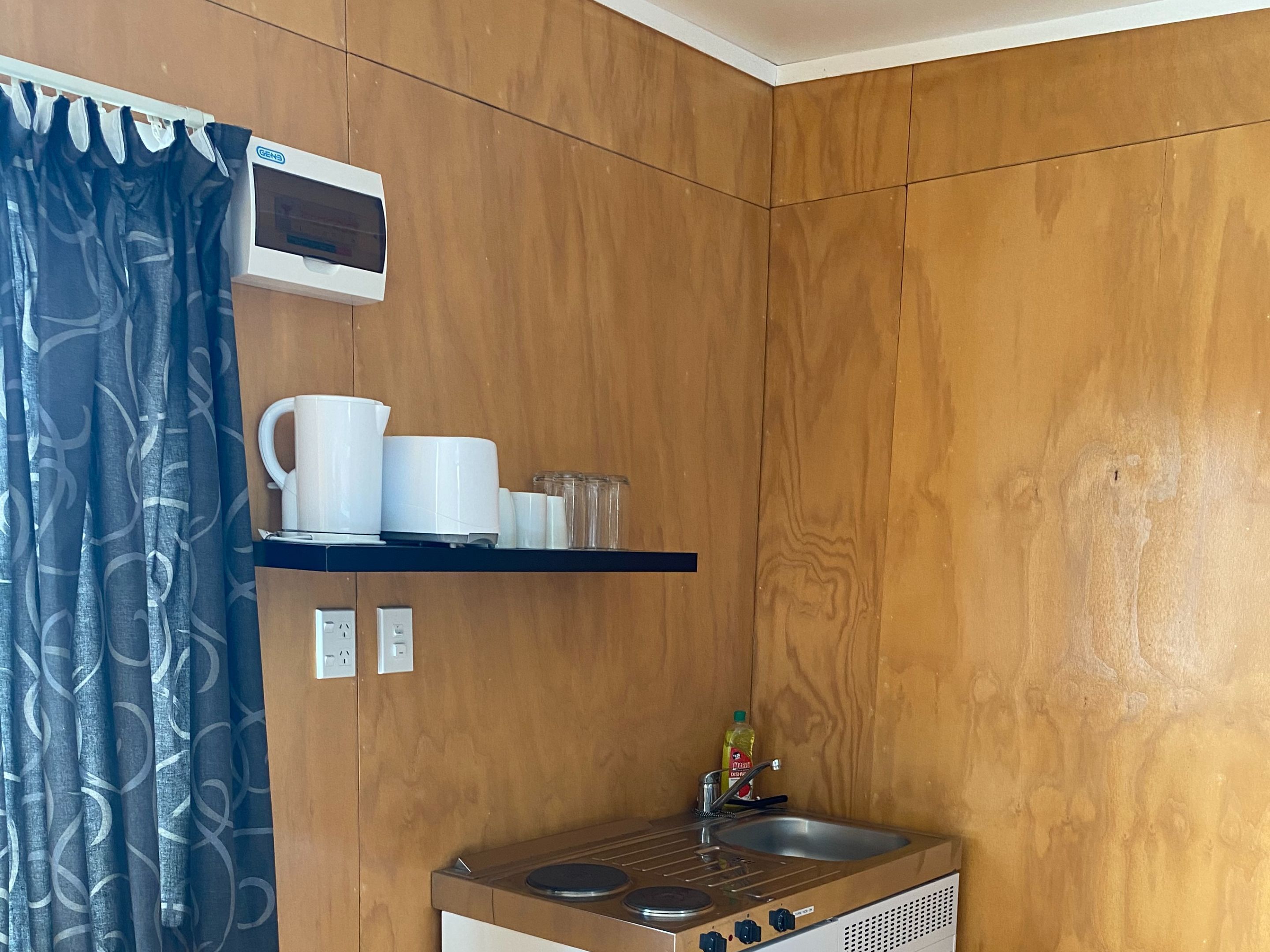 Una cocina compacta con estufa integrada, un lavamanos y un estante sobre la estufa con tazas blancas y un hervidor.