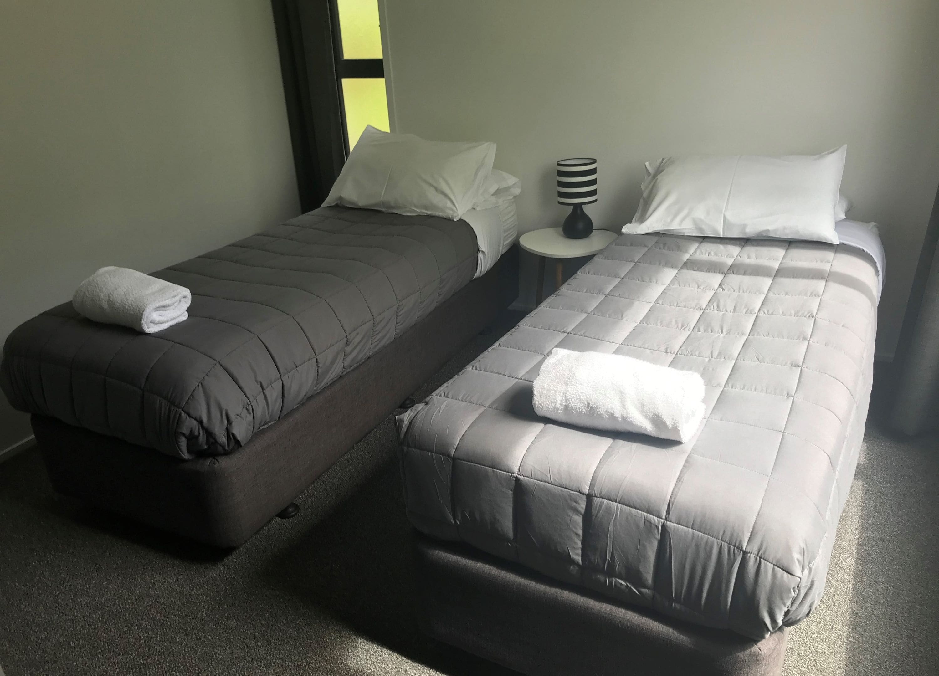A room with two single beds, each with a pillow and a folded towel. There is a bedside table with a lamp between the beds.