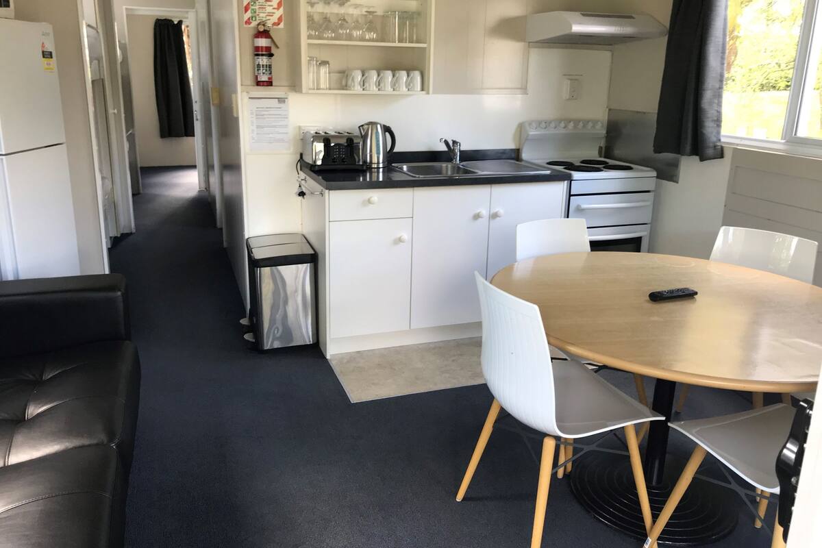 Une cuisine compacte avec des meubles blancs, un évier en acier inoxydable et un plan de travail noir. On y trouve une petite poubelle et une table à manger en bois, accompagnée de deux chaises.