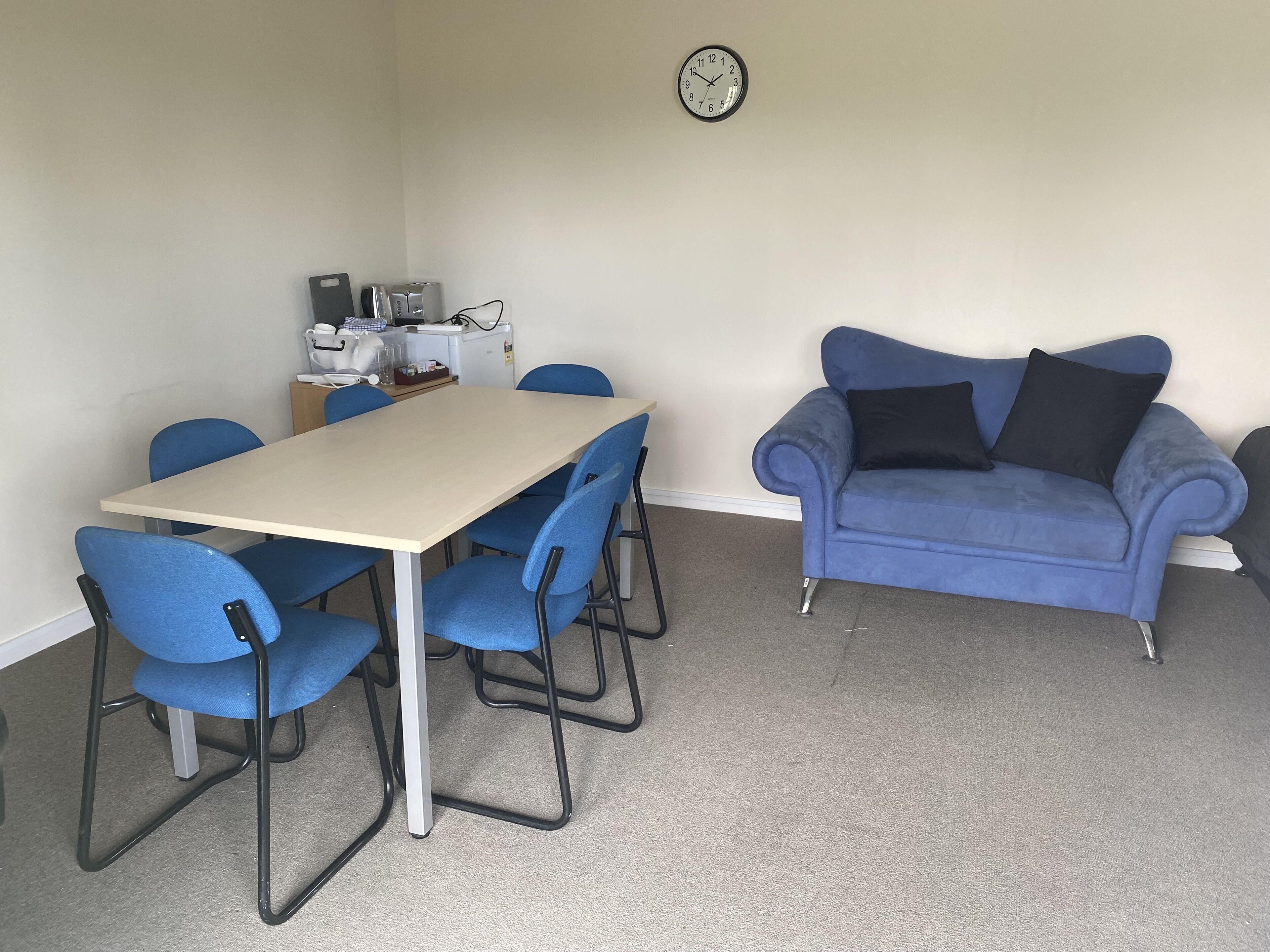 Una habitación con una mesa beige y sillas azules, un sofá azul con cojines negros, y un reloj en la pared.