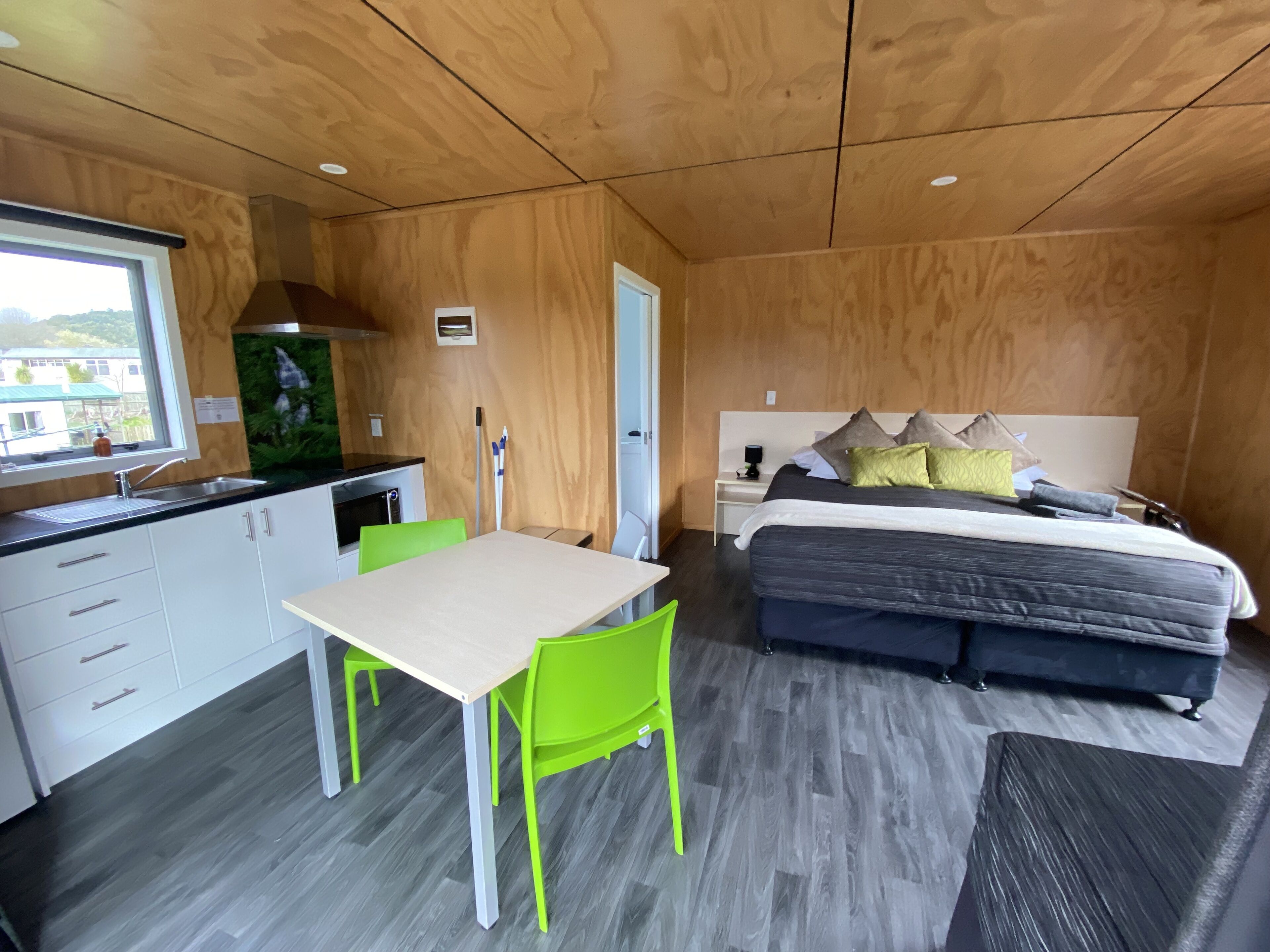 A modern room with a wooden ceiling, a bed with multiple pillows, a small table with chairs, a kitchenette, and a window with a view.