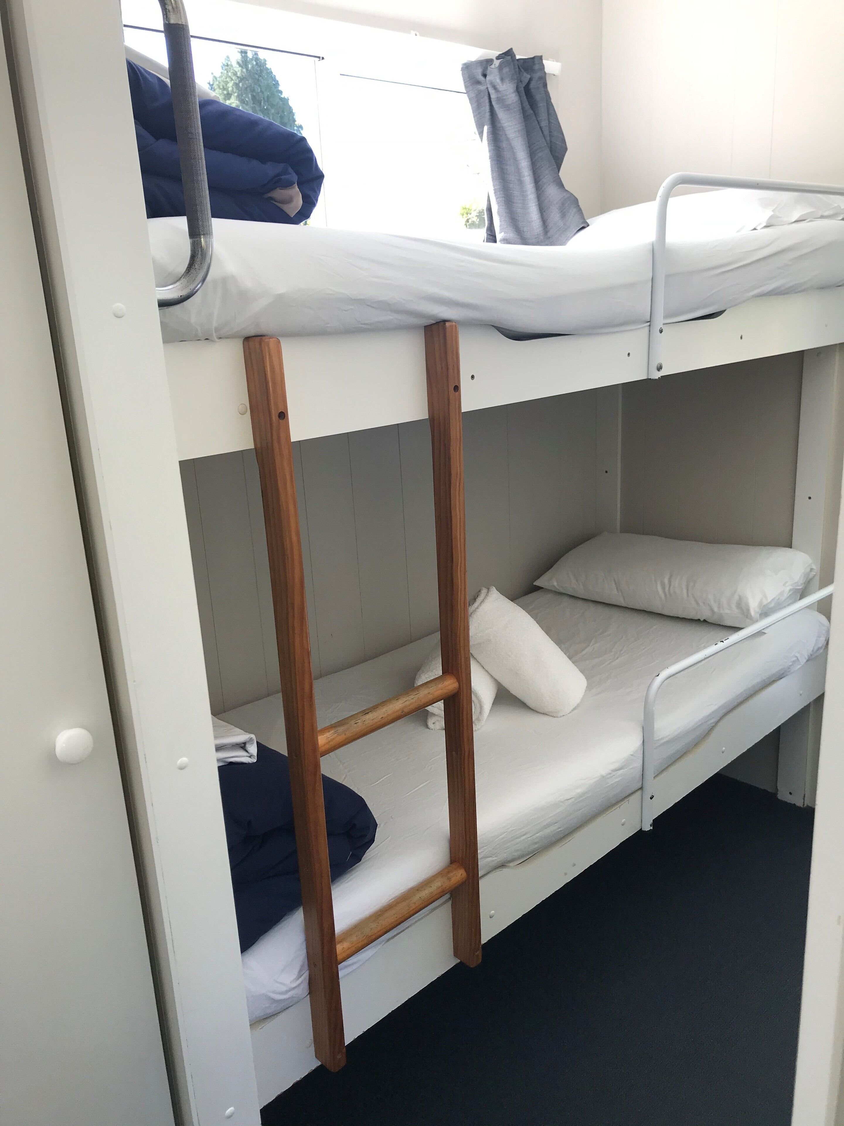 Un lit superposé avec une échelle en bois, du linge de lit blanc et une fenêtre avec des rideaux.