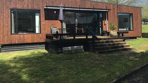 Una casa moderna de madera con amplios ventanales, un porche cubierto con mobiliario exterior y una escalera que conduce a la entrada.