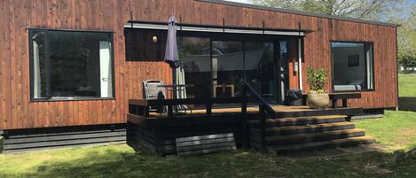 A modern wooden house with large windows, a covered patio with outdoor furniture, and a staircase leading to the entrance.