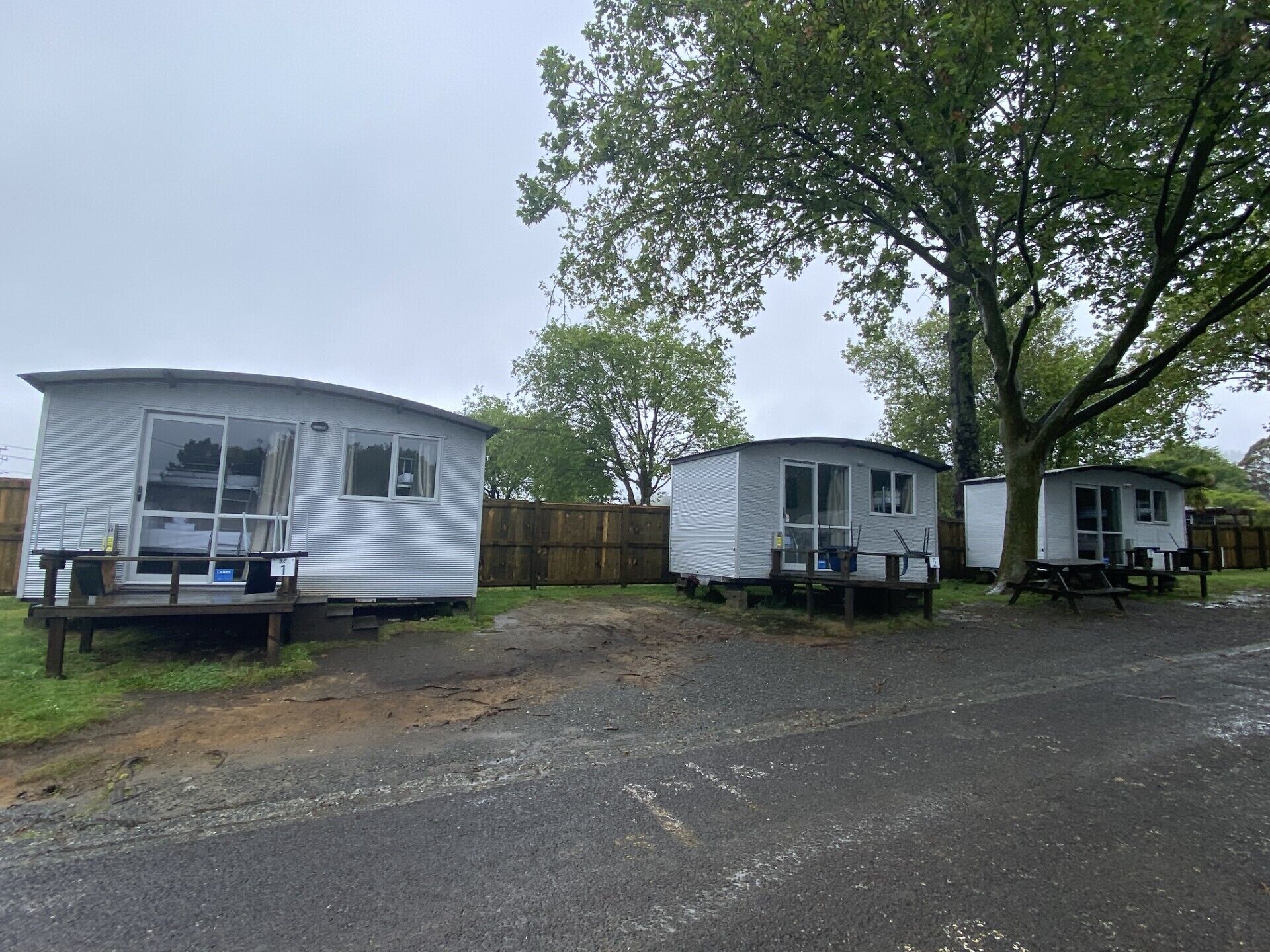 Two mobile homes with attached porches, a wooden fence, and a tree in the background.