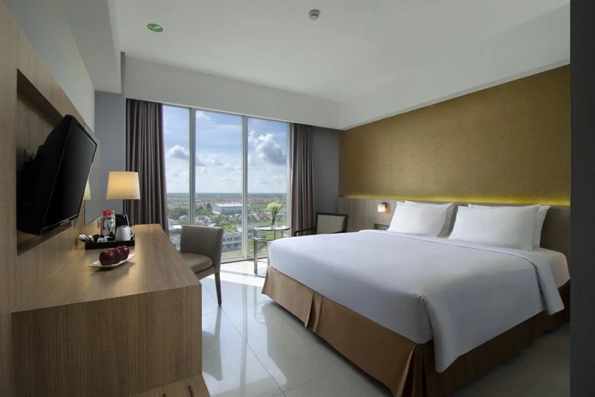Quarto de hotel com uma cama grande, uma escrivaninha com televisão, uma mesinha de apoio com abajur e vista para a cidade através de amplas janelas.