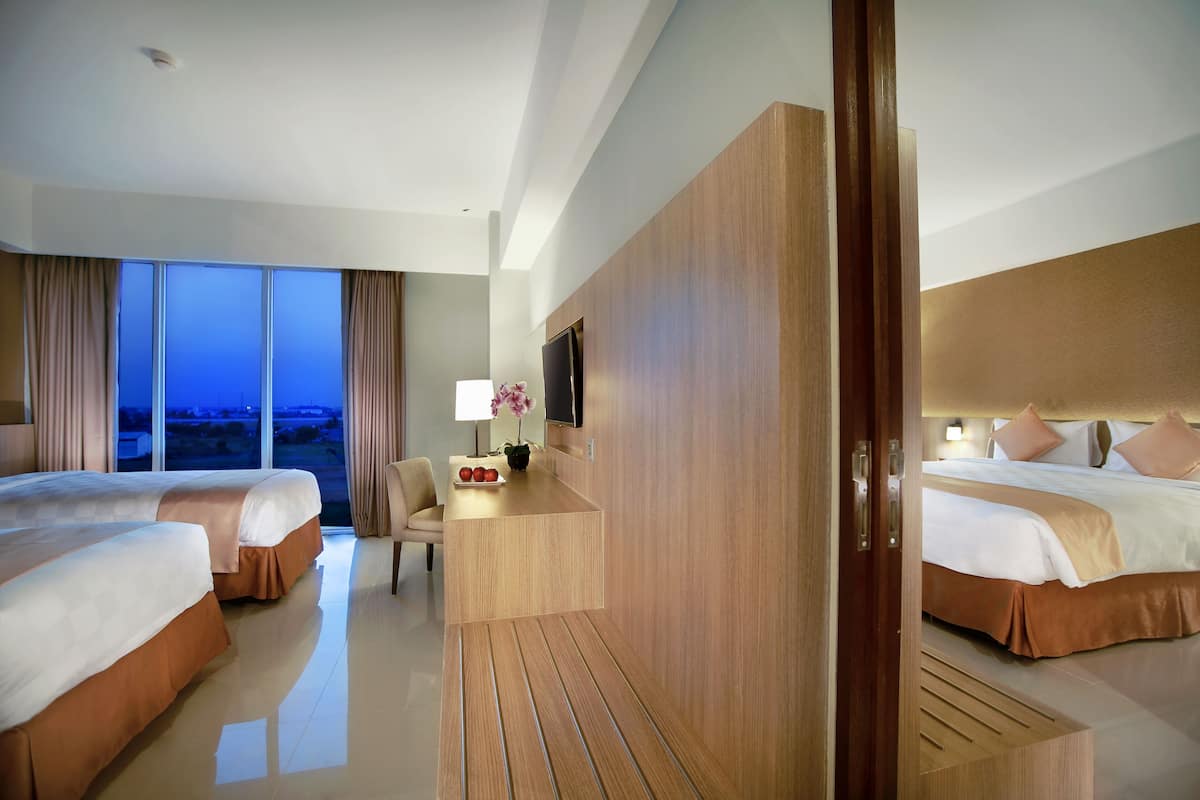Ein Hotelzimmer mit zwei Betten, einem Schreibtisch mit Lampe, einem Fernseher und einem großen Fenster mit Vorhängen.