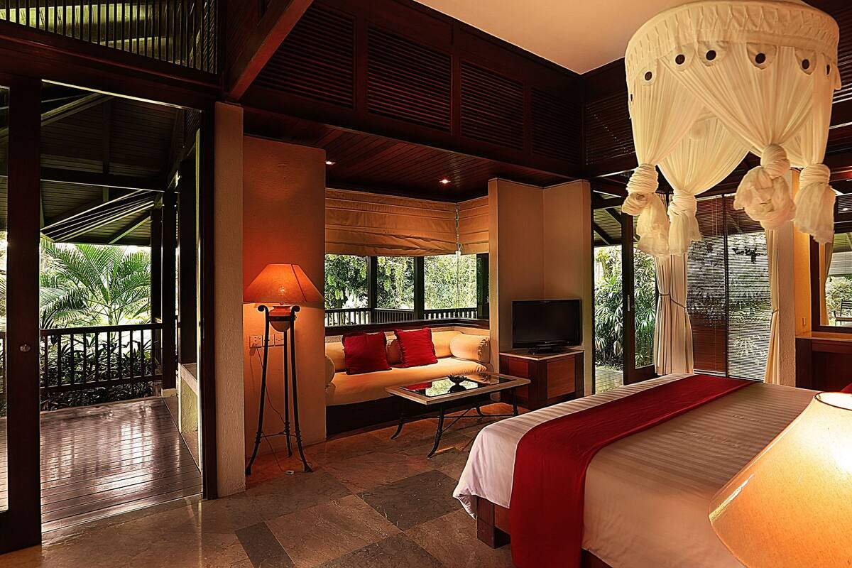 A bedroom with a canopy bed, a sofa, a TV, and a balcony with a view of greenery.