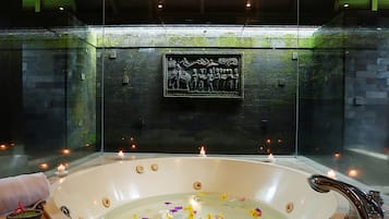 Un hidromasaje grande y ovalado con borde de madera, rodeado de velas, y una pared de piedra con un relieve escultórico.