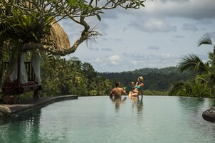 Una pareja relajándose en una piscina infinita privada, con un pabellón de techo de paja y abundante vegetación al fondo.