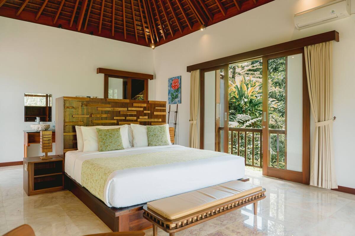 A bedroom with a large bed, a wooden headboard, a bedside table, and a view of greenery through a sliding door.