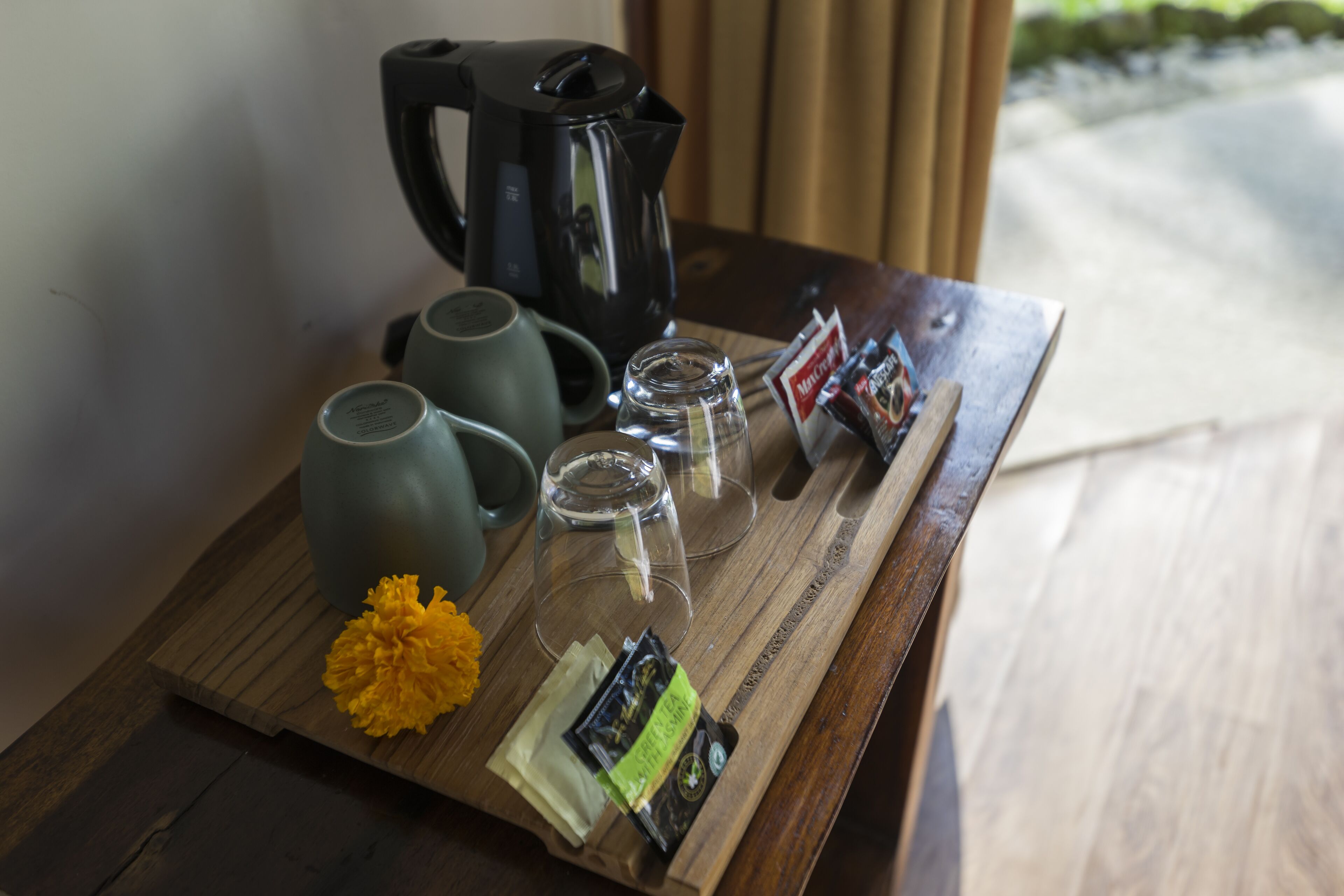 A wooden tray with a coffee maker, two mugs, a glass, and some snacks on a table.