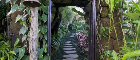 A stone pathway leading through a lush garden with a bell hanging from a tree.