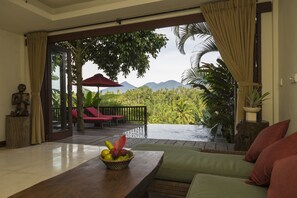 A room with a wooden table, a green sofa, and a view of a tropical landscape with palm trees and mountains. - Adiwana Dara Ayu (Payangan)
