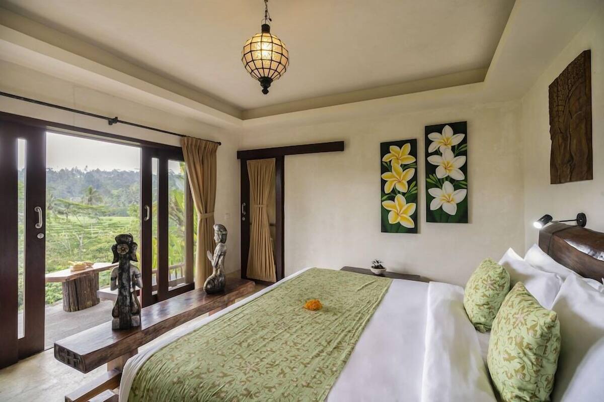 A bedroom with a bed, a view of the outdoors, a painting of flowers, and a statue.