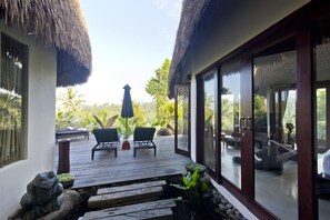 A wooden deck with two lounge chairs and a table, overlooking a tropical landscape with palm trees and a thatched-roof structure. - Adiwana Dara Ayu (Payangan)