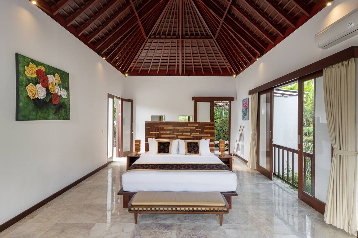 A spacious bedroom with a large bed, wooden ceiling, and a view of greenery through the windows.