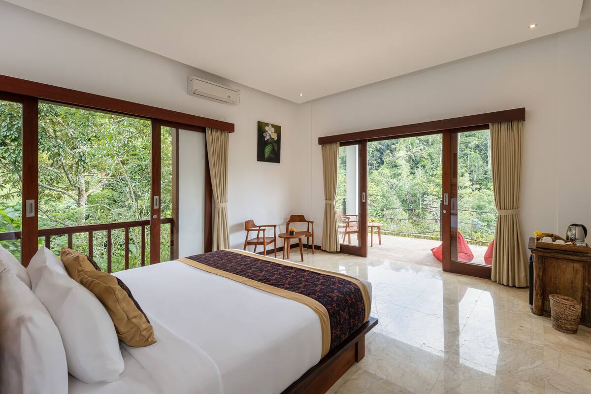A spacious bedroom with a large bed, a balcony with a table and chairs, and a view of the surrounding greenery.