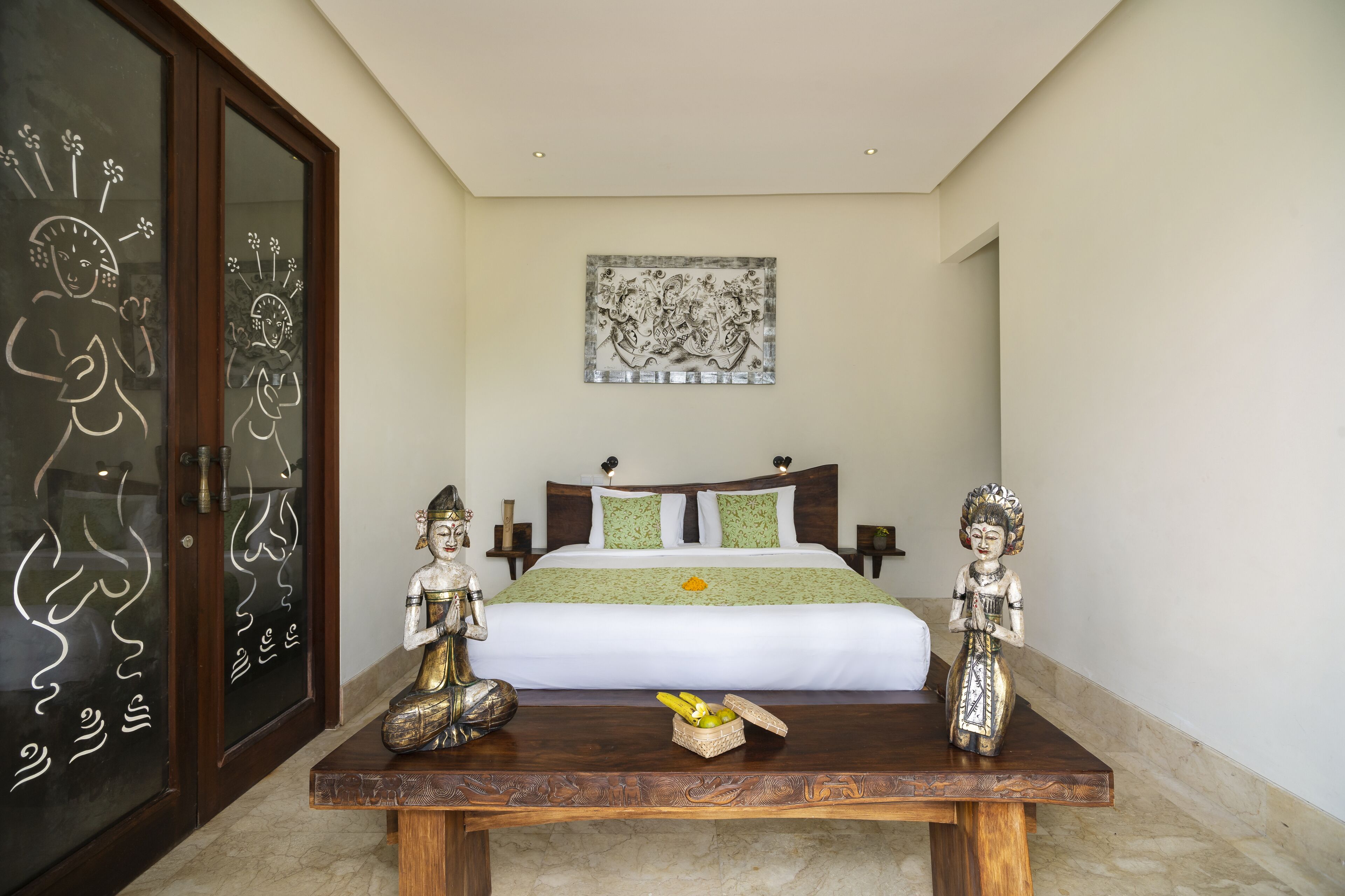 A bedroom with a bed, decorative statues, a table with fruit, and a wall with artwork.