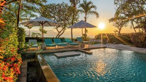 A pool area with lounge chairs and umbrellas, surrounded by tropical plants and palm trees, with the sun setting in the background.