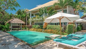 A pool area with lounge chairs and umbrellas, surrounded by lush greenery and a building with a tiled roof.