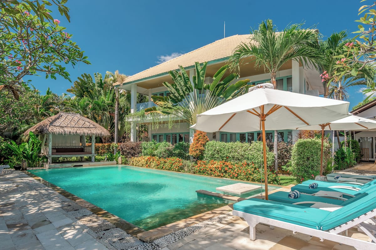 A pool area with lounge chairs and umbrellas, surrounded by lush greenery and a building with a tiled roof.