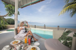 A person relaxing by a poolside with a view of the ocean, a dining setup with plates and drinks, and a tropical setting. - Mayo Resort (Seririt)