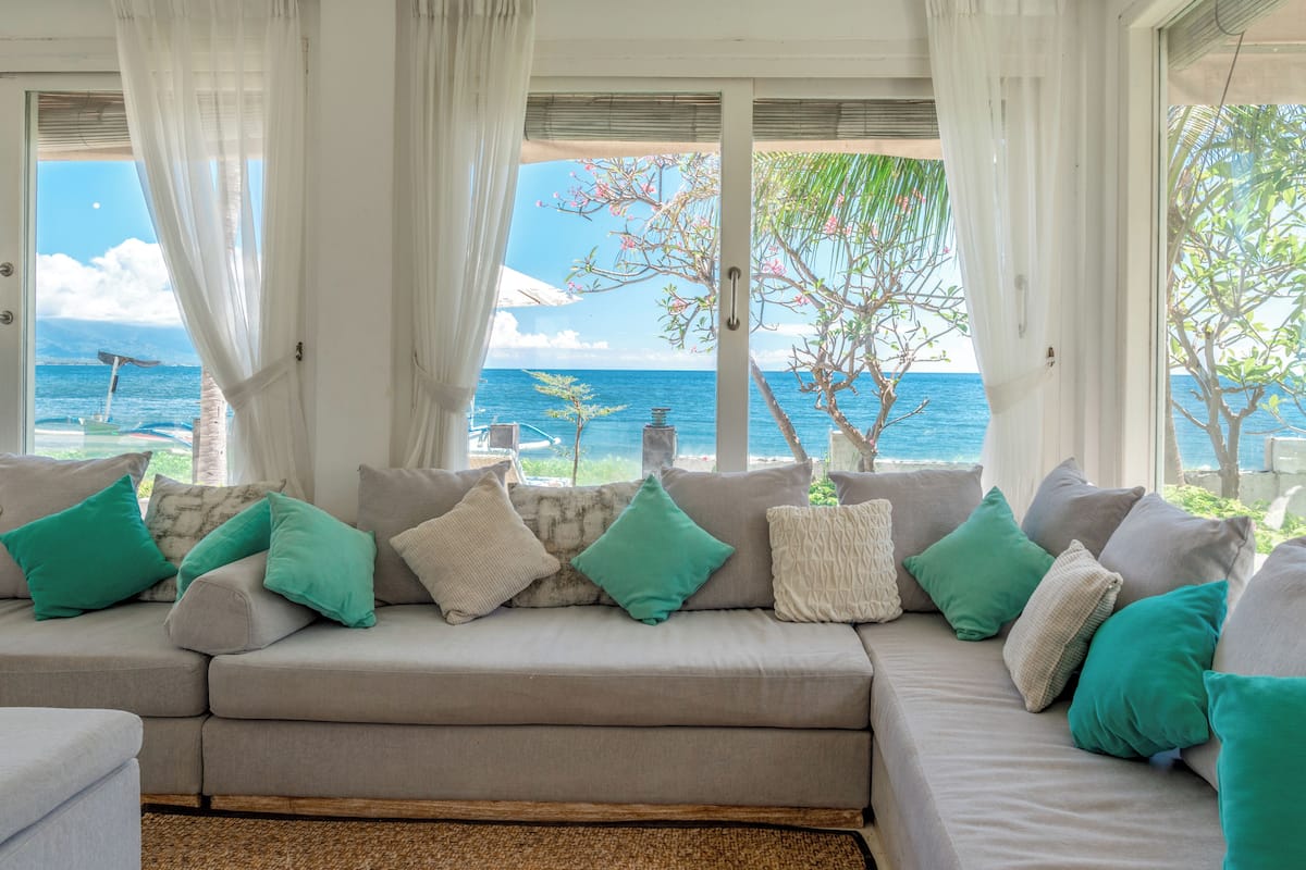 A beige sectional sofa with teal and white cushions, positioned in a room with large windows overlooking a beach and ocean.
