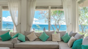 Un sofá seccional color beige con cojines verde azulado y blancos, ubicado en una habitación con ventanas amplias que dan a una playa y al océano.