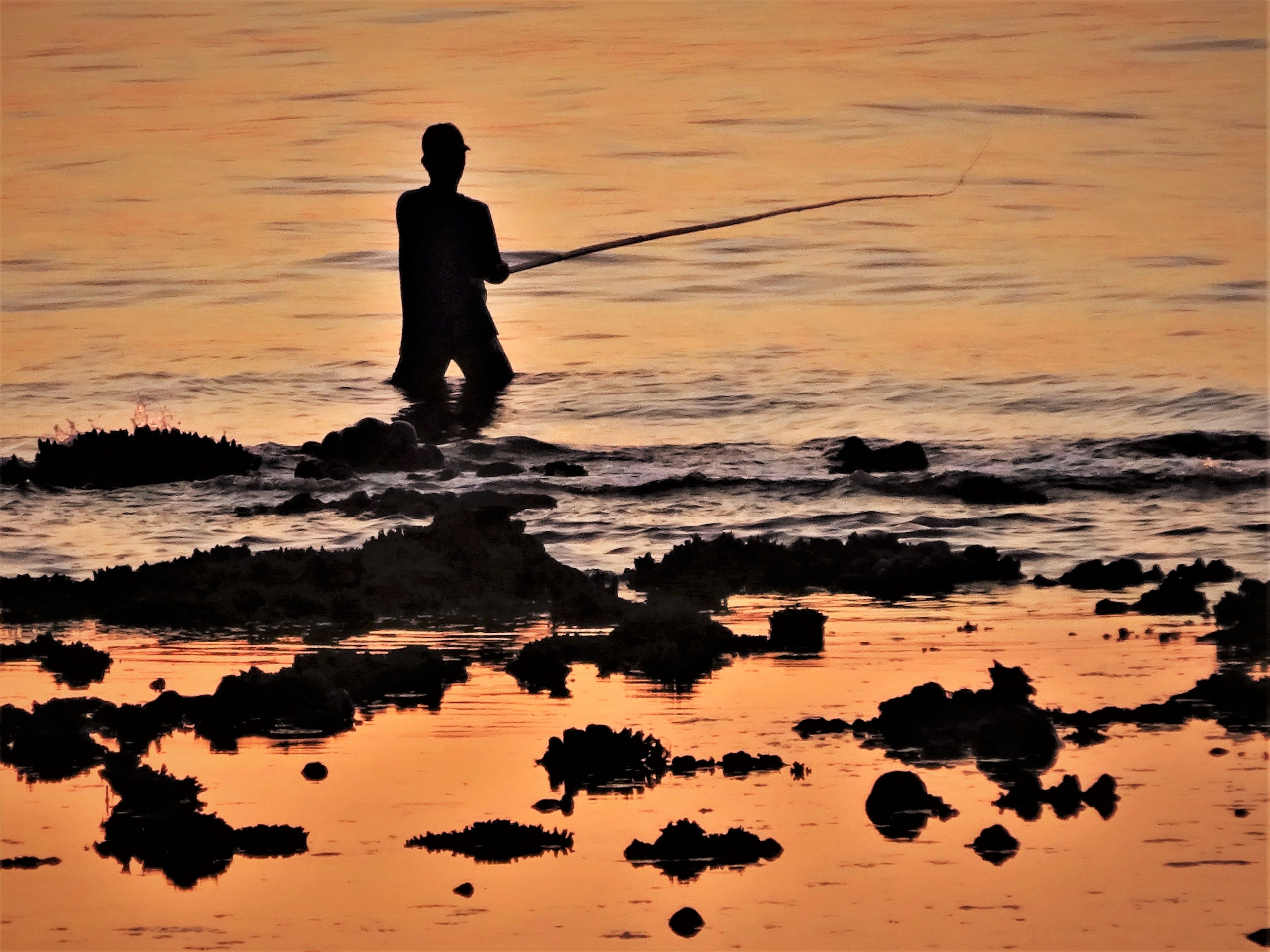 A silhouette of a person fishing at sunset with rocks in the foreground.