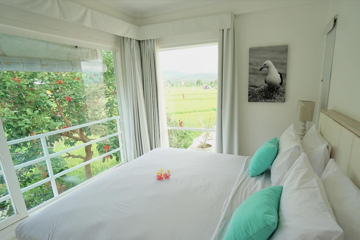 A bedroom with a large bed, a view of greenery outside, and a wall-mounted lamp.