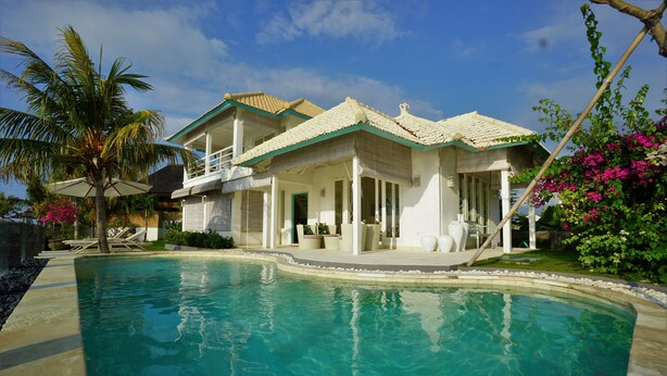 A poolside view of a house with a pool, a balcony, and a garden with palm trees and flowers.