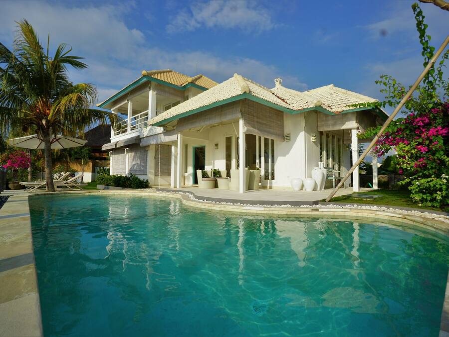 A poolside view of a house with a pool, a balcony, and a garden with palm trees and flowers.