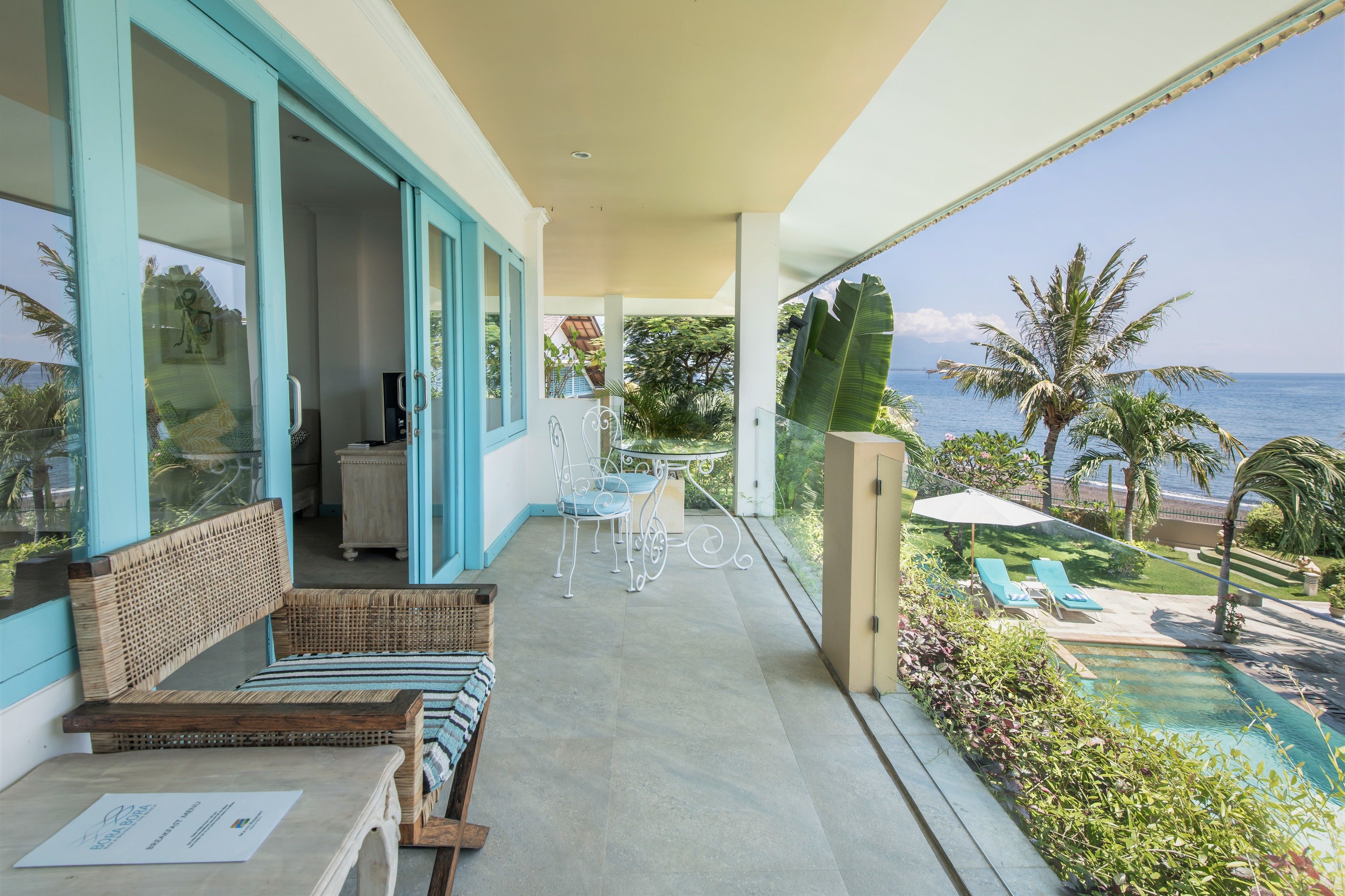 A balcony with a view of the ocean, a pool, and a dining area with outdoor furniture.