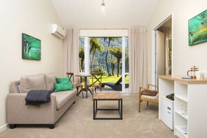Uma sala de estar com sofá bege, uma mesa de centro, uma mesa de jantar com cadeiras e vista para uma paisagem tropical através de uma porta de vidro.