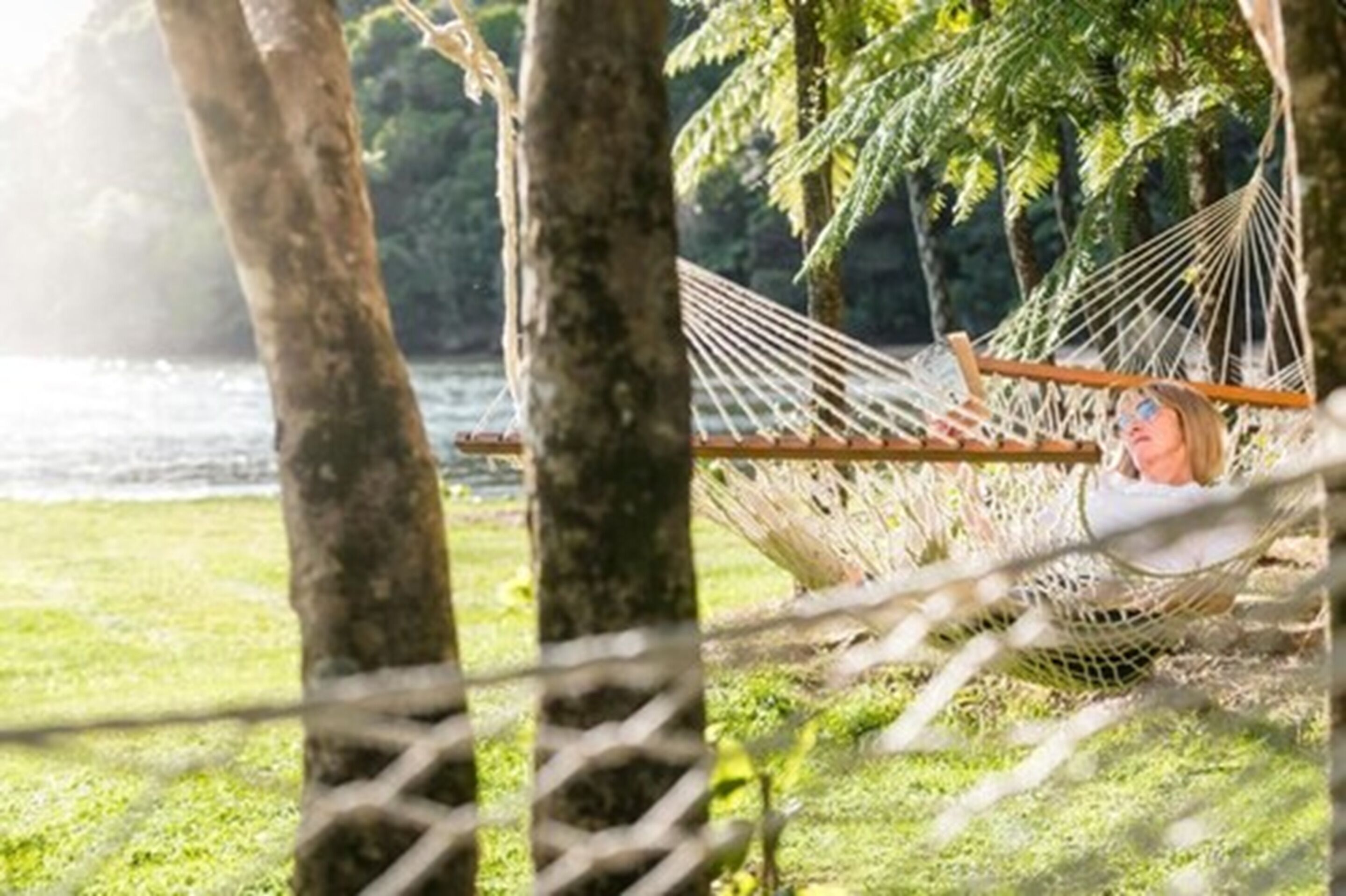 A hammock tied between trees in a grassy area with a body of water in the background.